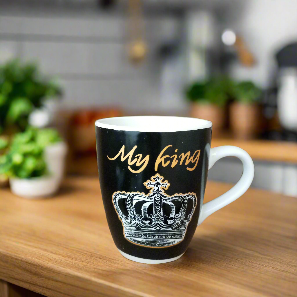 A black ceramic mug with 'My King' text and a crown design on it, placed on a wooden surface with a blurred kitchen background.
