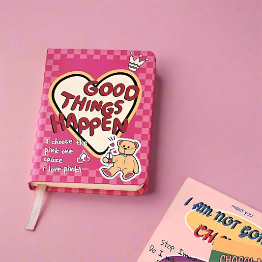 Pink notebook with 'Good Things Happen' design on a white surface