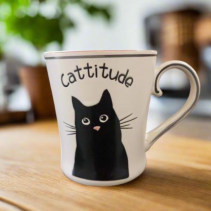 A white ceramic mug with a black cat face illustration and the word 'Cattitude' written on it.