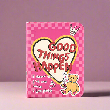 Pink checkered notebook with 'Good Things Happen' text and cartoon bear on a white background